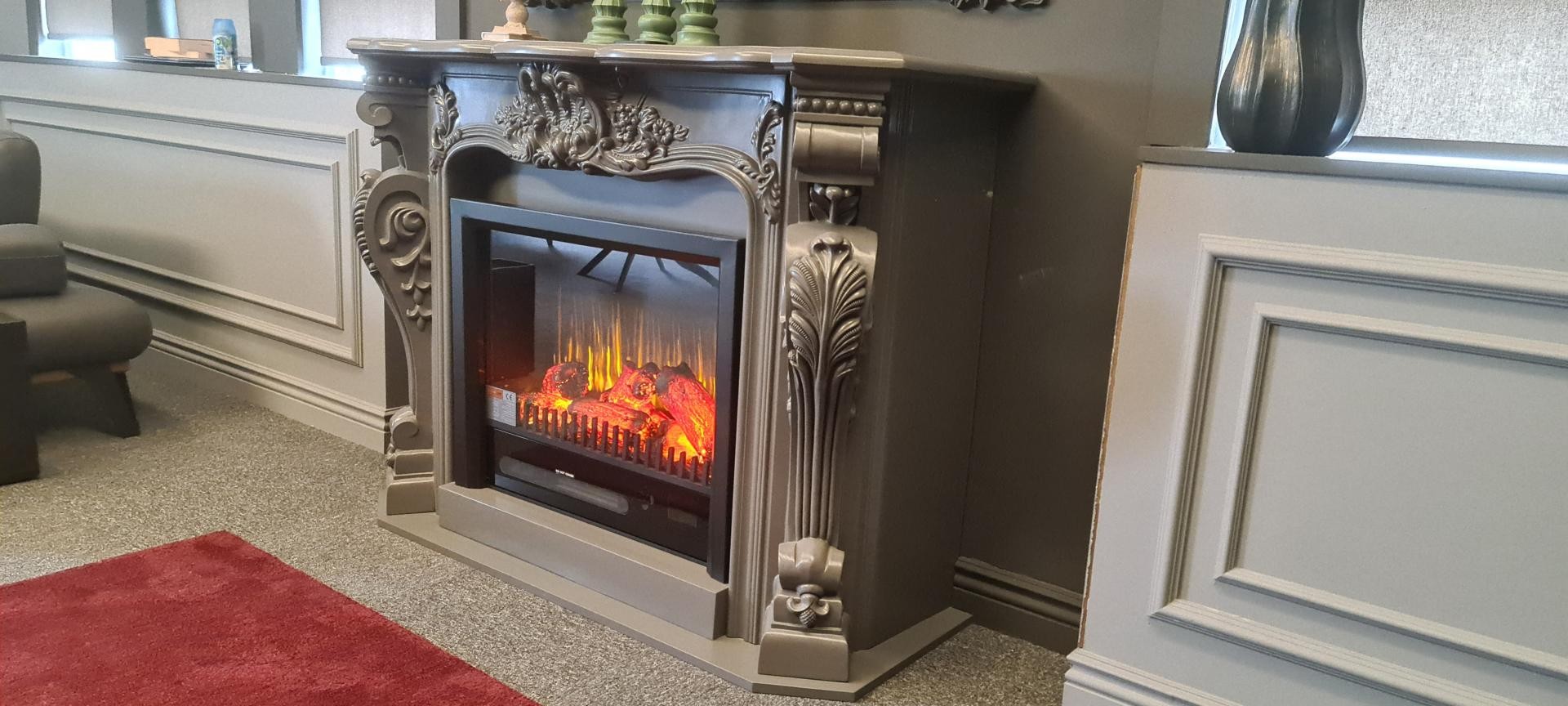 Semișemineu gri-antracit cu foc electric și console sculptate, pe covor roșu în showroom