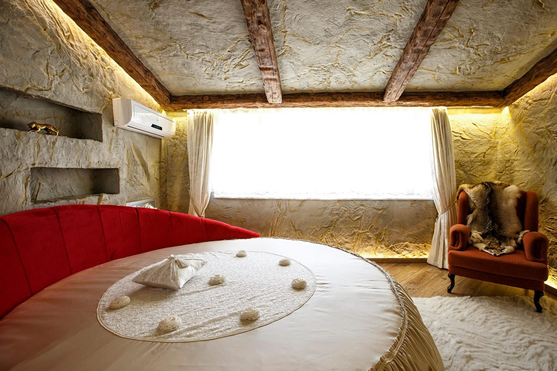 Dormitor rustic cu grinzi decorative aparente, pereți texturați și pat rotund cu canapea roșie