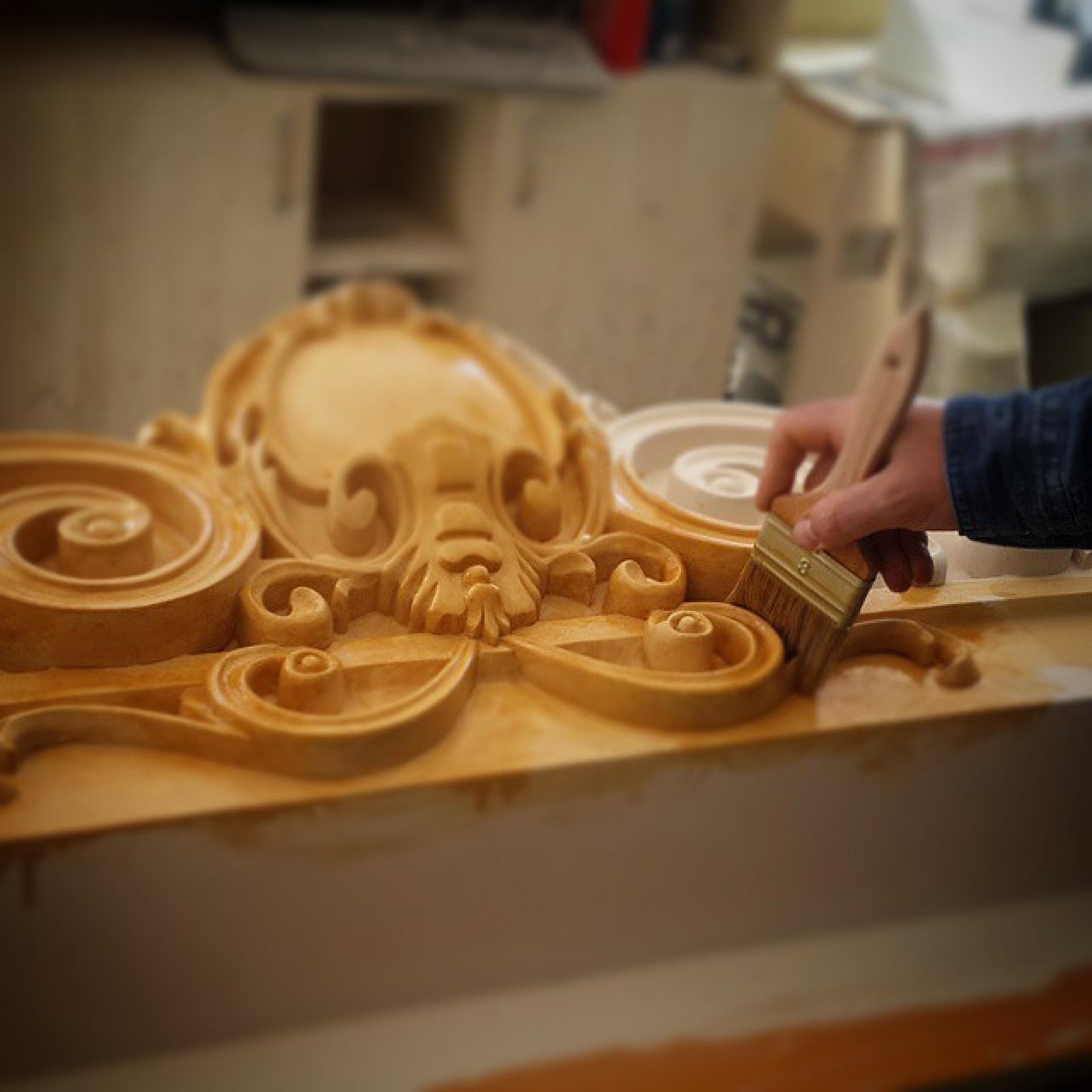 Mână aplicând lac cu pensula pe un model decorativ cu volute și cartuș oval, în atelier