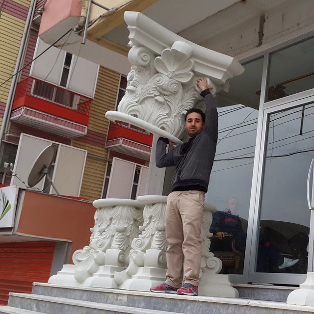 Bărbat ridicând un capitel corintic alb din poliuretan lângă o coloană, pe stradă