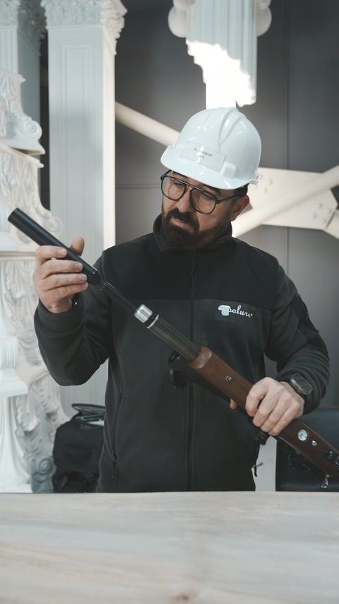 Muncitorul examinează arma de testare deasupra grinzii cu textură lemnoasă.